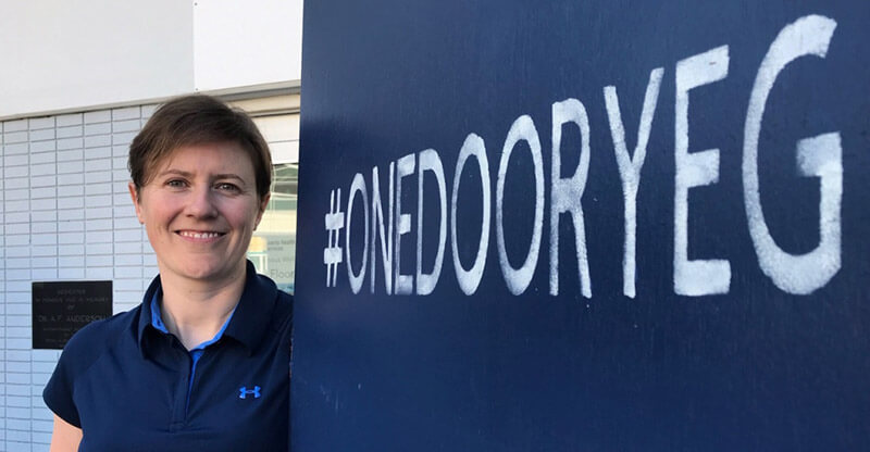 A woman with short brown hair stands by a sign with the hashtag #OneDoorYEG.
