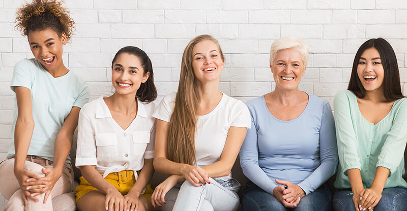 A group of women of various ages and race sit together smiling at the camera.