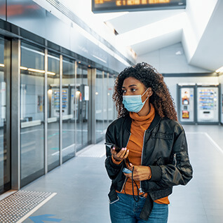 A young woman is walking through a public transit terminal, she is wearing a surgical mask.
