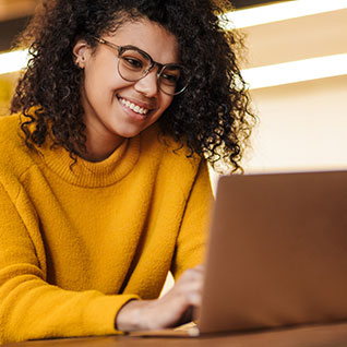A young woman is at a desk, looking at a laptop. She is smiling.