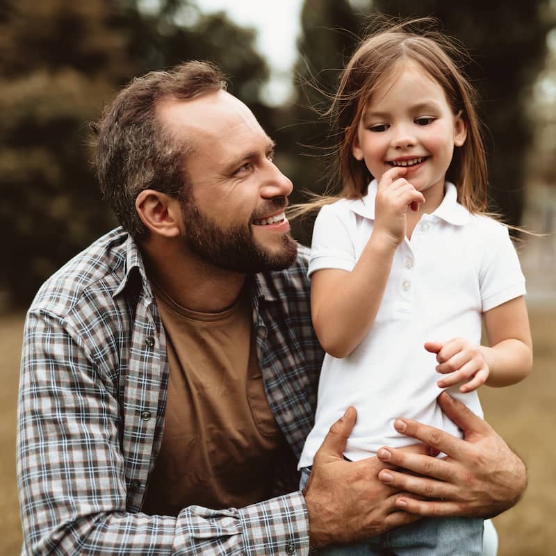 A young Caucasian father knees on the ground next to this daughter. He is looking at her face and smiling.