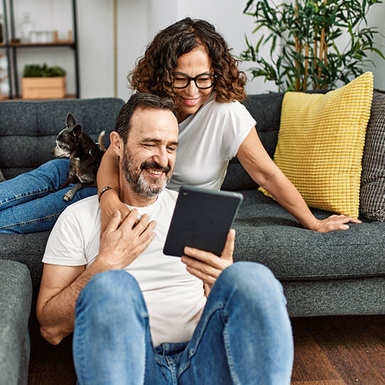 A happy Hispanic couple are looking at a tablet together. The woman is stretch out on a couch behind the man, who is sitting on the floor. There is a small dog in the woman's lap.