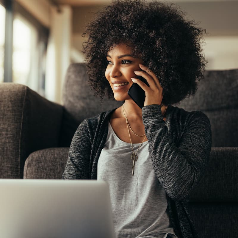 A young African-Canadian woman is sitting against a couch, with her laptop on her lap. She is holding her smartphone up to her eye and smiling as she looks off to the left.