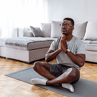 A young man siting cross-legged on a yoga mat in a naturally-lit room with his eyes closed and hands in a prayer.