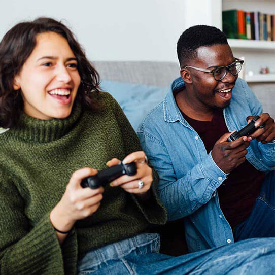 Two friends are sitting on a couch, they are holding a Play Station controllers and smiling.