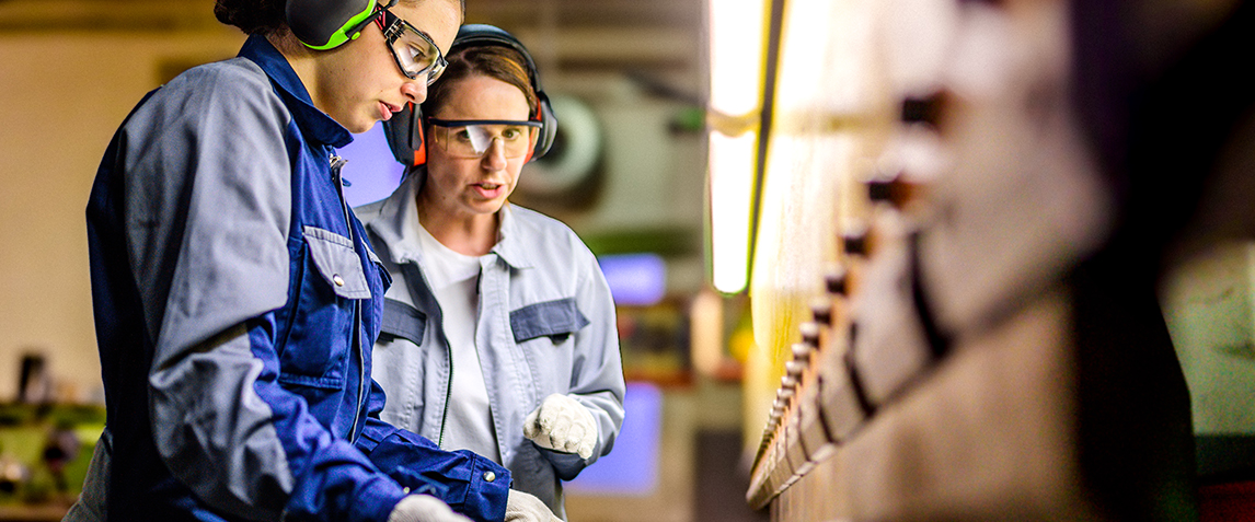Two women working in with heavy equipment wearing safety gear.