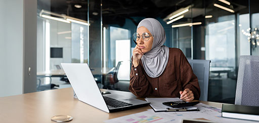 A wman wearing glasses sitting at a desk and looking at a laptop with a calculator and clipboard on her desk nearby.