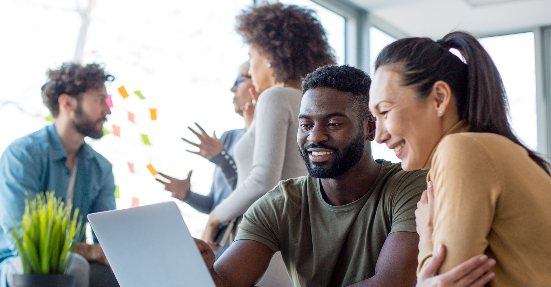 Coworkers smiling and working together on a laptop, with a team discussing in the background.