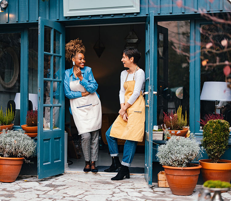 Two people wearing aprons tanding in the doorway of blue French doors while talking and smiling.