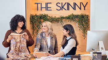 Founder of The Skinny posing in the store with 2 workers folding clothes.