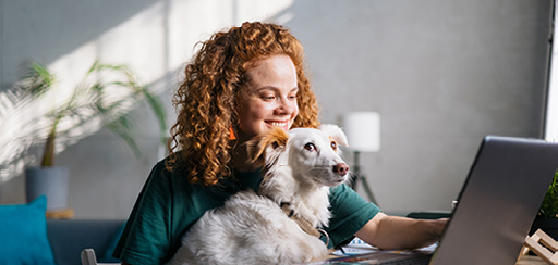 A red hair girl smiling at her computer with her dog on her lap.